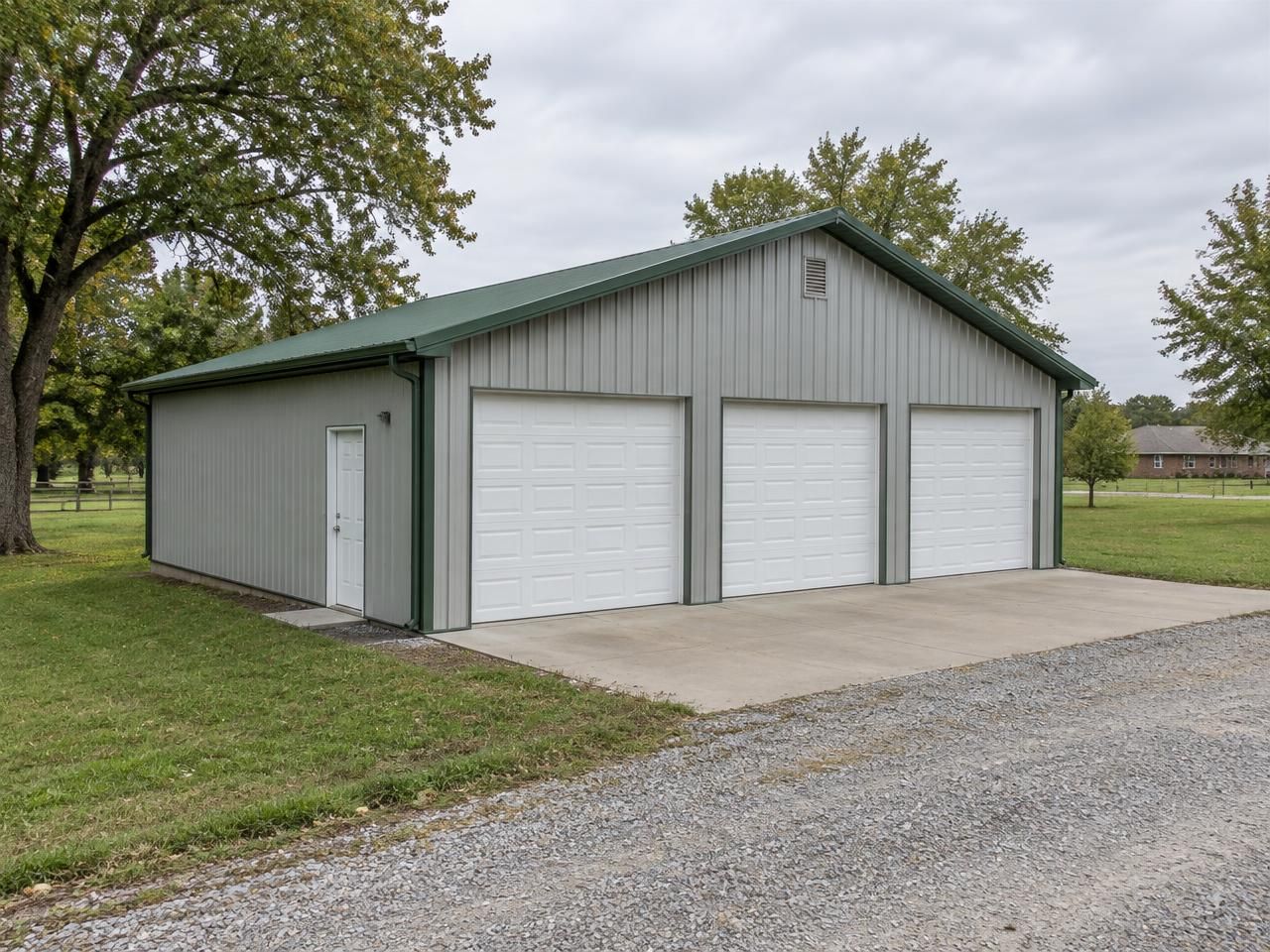 Illustrative building concept — AI-generated, not a completed Outlaw Building Co. project AI-generated illustration: Detached light-gray metal garage with three overhead doors and green roof trim on an Oklahoma-style rural property. Not a completed Outlaw Building Co. project.