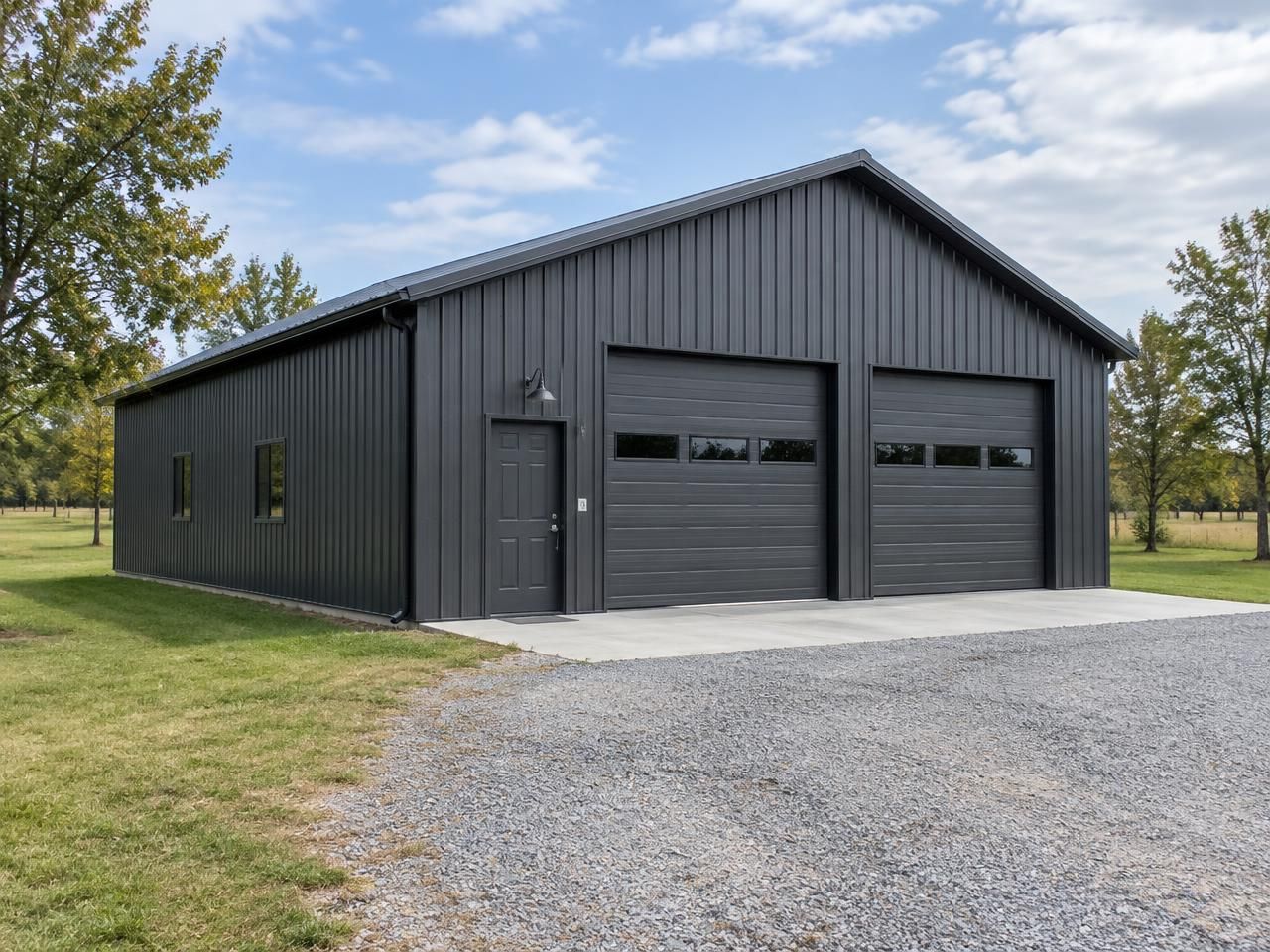 Illustrative building concept — AI-generated, not a completed Outlaw Building Co. project AI-generated illustration: Charcoal metal workshop with two overhead doors, a concrete apron and gravel driveway in daylight. Not a completed Outlaw Building Co. project.